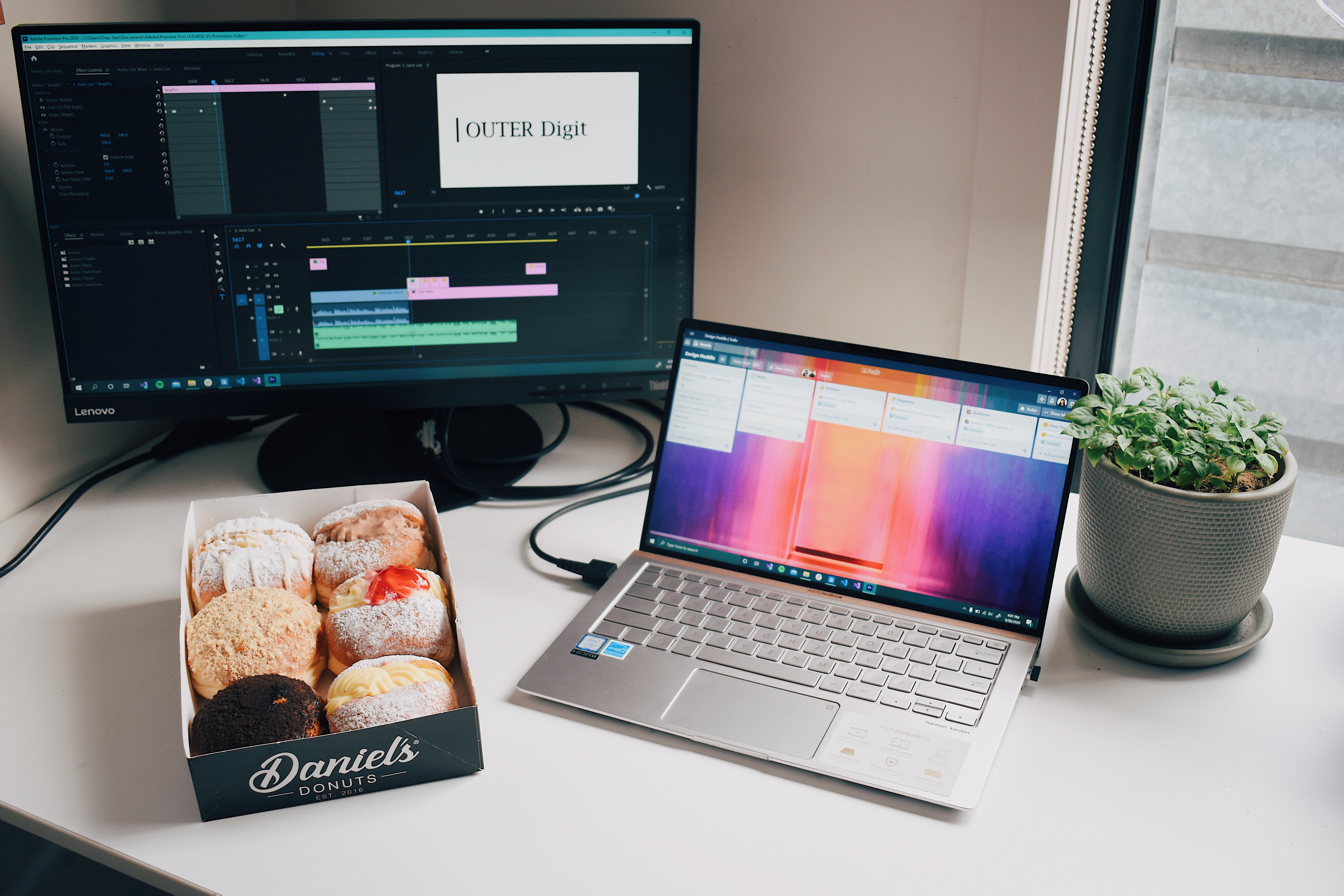 An office workspace with a laptop, a monitor, a plant, and a box of donuts.